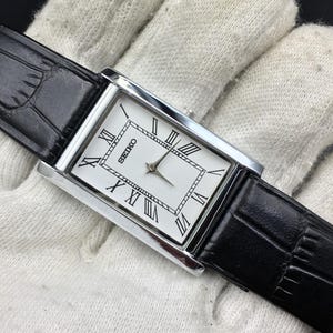 May include: A rectangular Seiko wristwatch with a silver-tone case and a white dial featuring black Roman numeral hour markers. The watch has a black leather strap with a textured finish and silver-tone hands for the hour and minute display.