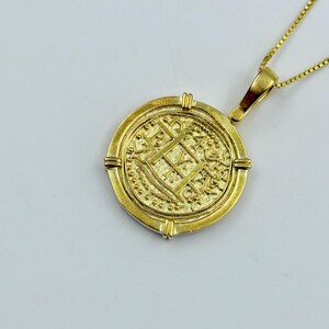 May include: A gold-colored coin pendant necklace. The round pendant features intricate embossed designs and is suspended from a delicate gold chain. The coin has a detailed pattern with a cross and other symbols.