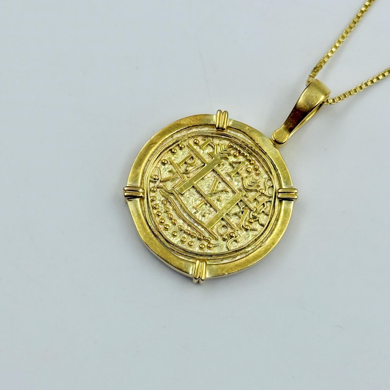 May include: A gold-colored coin pendant necklace. The round pendant features intricate embossed designs and is suspended from a delicate gold chain. The coin has a detailed pattern with a cross and other symbols.