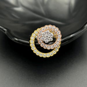 May include: A multi-tone ring featuring a central cluster of small, clear stones surrounded by two concentric circles. The outer circle is gold-toned, the middle is rose gold-toned, and the center is silver-toned, all encrusted with clear stones. The ring is set against a dark background.