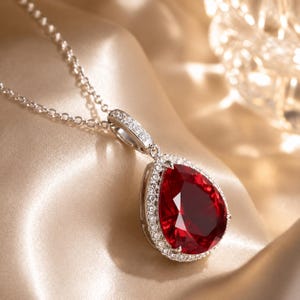 May include: A silver-toned necklace featuring a large, pear-shaped, deep red gemstone pendant. The pendant is surrounded by a halo of small, clear, sparkling stones, with additional clear stones set along the bail connecting to a delicate chain.