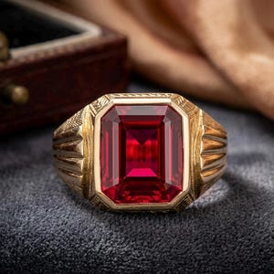 Puede incluir: Un anillo de oro con una gran piedra preciosa roja de corte esmeralda rectangular en el centro. La banda de oro muestra detalles intrincados, texturizados y ranurados a los lados, enmarcando la piedra roja vibrante con un acabado pulido.