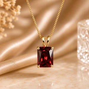 May include: A gold necklace featuring a rectangular, deep red gemstone pendant held by four prongs. The pendant is suspended from a delicate gold chain, showcasing a faceted cut that reflects light across the surface of the dark red stone.