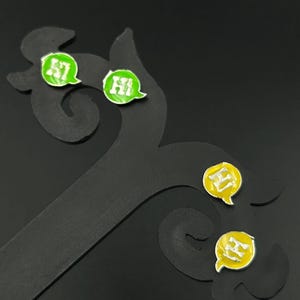 Könnte beinhalten: Ohrringe mit Sprechblasen-Design. Die Ohrringe haben die Form von Sprechblasen mit dem Wort "Hi" in weißen Buchstaben. Sie sind grün und gelb mit einem silbernen Rand. Die Ohrringe werden auf einem schwarzen Ständer präsentiert.