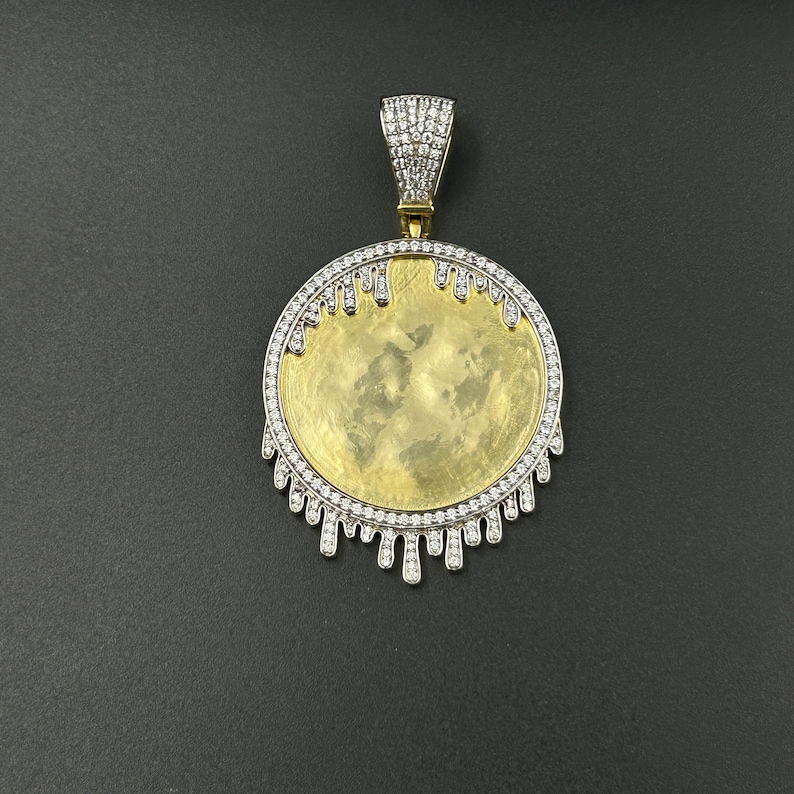 Op de afbeelding: Een goudkleurige hanger met een cirkelvormig ontwerp. De hanger heeft een goudkleurig midden, een zilverkleurige rand bezet met kleine, heldere stenen en een decoratieve, druipende rand. De beugel is ook bezet met kleine, heldere stenen. De hanger wordt tegen een donkere achtergrond gepresenteerd.
