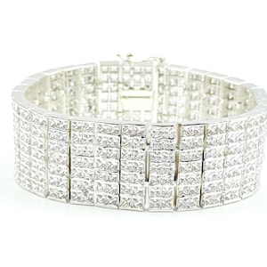 Puede incluir: Un brazalete ancho, plateado, con múltiples filas de gemas cuadradas y transparentes. El brazalete tiene un cierre seguro y está sobre un fondo blanco. Las gemas reflejan la luz, creando un efecto brillante. Esta pieza de joyería es adecuada para ocasiones especiales.