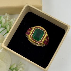 Puede incluir: Un anillo de oro con una gran piedra verde de talla esmeralda en el centro, rodeada de piedras rojas más pequeñas. El anillo está engastado en una banda de oro y se muestra en una pequeña caja abierta con interior negro.