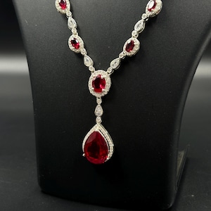 May include: A silver-toned necklace with a series of oval and teardrop-shaped red gemstones, each surrounded by smaller, clear stones. The pendant features a large, pear-shaped red gemstone. The necklace is displayed on a black stand.