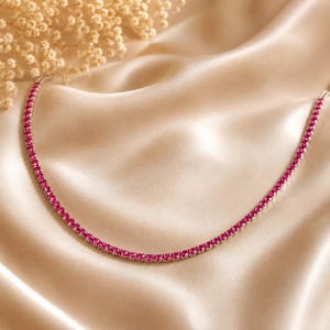 Könnte beinhalten: Eine Tennis-Halskette mit einer durchgehenden Reihe runder, leuchtend pinker Edelsteine, die in eine polierte, silberfarbene Metallkette gefasst sind. Die Kette ist in einer sanften Kurve angeordnet und zeigt den einheitlichen Schliff der Steine.