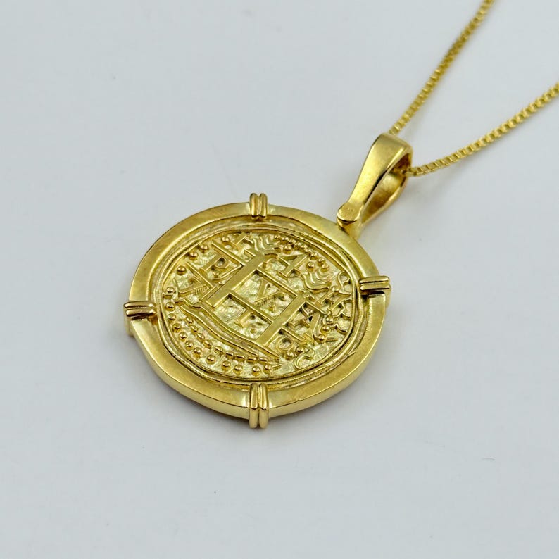 May include: A gold-colored pendant necklace featuring a round coin-like charm. The charm has intricate embossed details and is suspended from a delicate gold chain. The pendant has a rectangular design in the center.