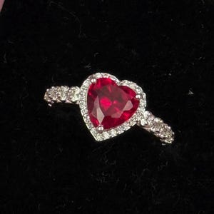 Puede incluir: Anillo en forma de corazón con una gran gema roja facetada en el centro. La gema está rodeada por un halo de piedras más pequeñas y transparentes. La banda es de color plateado y también presenta piedras transparentes.