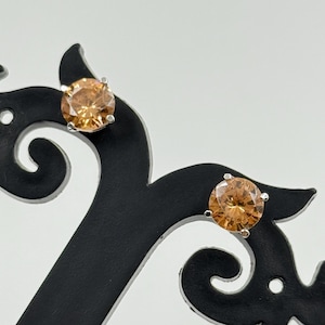 May include: A pair of stud earrings with round, light orange gemstones set in silver-coloured metal. The earrings are displayed on a black decorative stand against a white background. The gemstones have a faceted appearance.