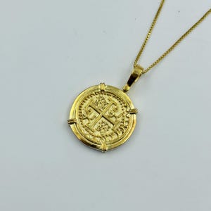 May include: A gold-colored coin pendant necklace. The pendant is round with an embossed design featuring a cross and ornate details. The necklace chain is a delicate gold color. The pendant is approximately 1 inch in diameter.