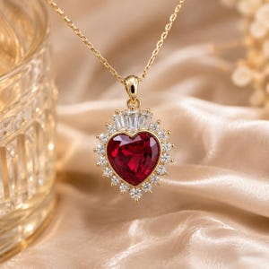Op de afbeelding: Een gouden ketting met een hartvormige rode edelsteen hanger, omringd door een halo van kleine ronde heldere stenen en bekroond met drie heldere stenen in baguette-slijpvorm. De hanger hangt aan een fijne gouden ketting.