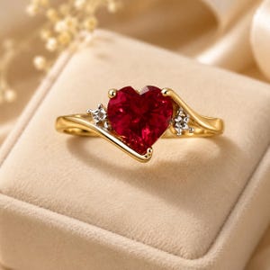 Könnte beinhalten: Ein Goldring mit einem zentralen herzförmigen roten Edelstein. Die Ringschiene hat ein geschwungenes Design mit zwei kleinen klaren Steinen, die auf beiden Seiten des Herzens gefasst sind. Das Metall ist poliertes Gelbgold mit weißer Fassung.