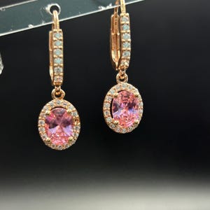 May include: Rose gold-tone earrings featuring oval pink gemstones surrounded by small, clear stones. The earrings have a lever back closure and are displayed against a dark background. The earrings are a classic jewelry design.