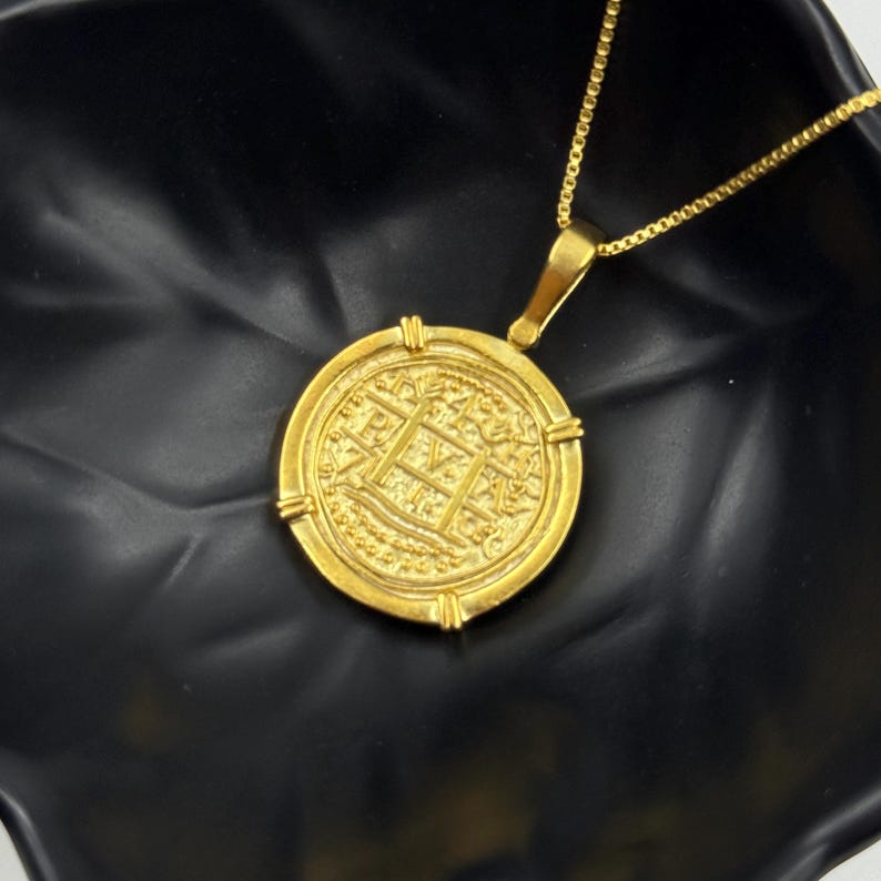May include: A gold-colored pendant necklace featuring a round coin-like charm. The charm has an intricate design with a central grid pattern and decorative elements around the edge. The necklace chain is also gold-colored.