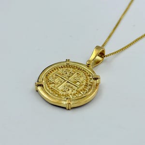 May include: A gold-colored pendant necklace featuring a round coin design. The coin has an embossed cross and intricate detailing. The necklace chain is also gold-colored, creating a cohesive and elegant look. The pendant is suspended from a delicate chain.