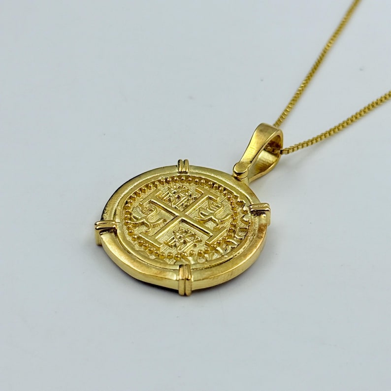 May include: A gold-colored pendant necklace featuring a round coin design. The coin has an embossed cross and intricate detailing. The necklace chain is also gold-colored, creating a cohesive and elegant look. The pendant is suspended from a delicate chain.