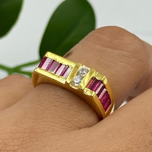 May include: A gold ring featuring a rectangular design with alternating sections of red gemstones and clear stones. The ring is set on a finger, showcasing its bright, polished finish. The gemstones are set in a channel setting.