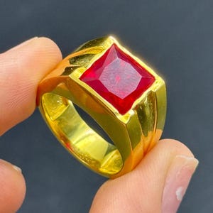 Puede incluir: Un anillo de oro con una gran gema roja brillante de talla cuadrada. El anillo tiene una banda gruesa y una superficie pulida y reflectante. La gema está engastada en un bisel de oro. El anillo se sostiene entre los dedos.
