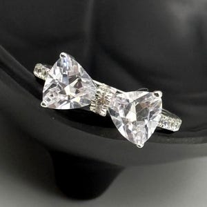May include: A silver-toned ring featuring a bow design. The bow is formed by two large, clear, triangular-cut stones. The band is embellished with smaller, clear stones. The ring is set against a black background.
