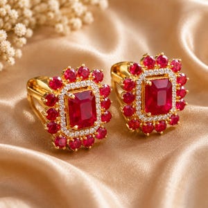 May include: A pair of gold earrings featuring a large rectangular red gemstone center, surrounded by a halo of small white stones and an outer border of round red stones. The design is symmetrical with a polished gold metal setting and backing.