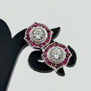May include: A pair of silver-toned stud earrings. Each earring features a large, round, clear center stone surrounded by smaller, square, deep pink stones. The earrings are displayed on a black stand against a white background.
