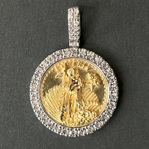 May include: A gold-colored pendant with a Liberty coin design, surrounded by a ring of small, clear stones. The bail is also encrusted with similar stones. The pendant is set against a dark background.
