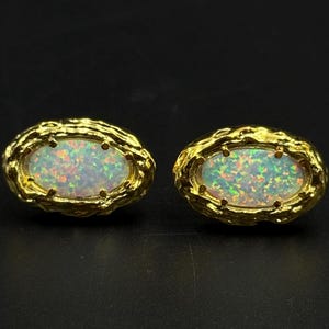 May include: A pair of gold-colored oval stud earrings. Each earring features a central opal stone with iridescent colors, set within a textured gold frame. The earrings are displayed against a dark background.