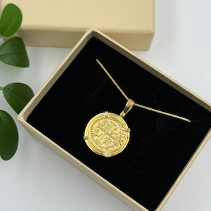 May include: A gold-colored coin pendant necklace displayed in a gift box. The pendant is round with an embossed design and is suspended from a delicate gold chain. The box is beige with a black velvet interior.