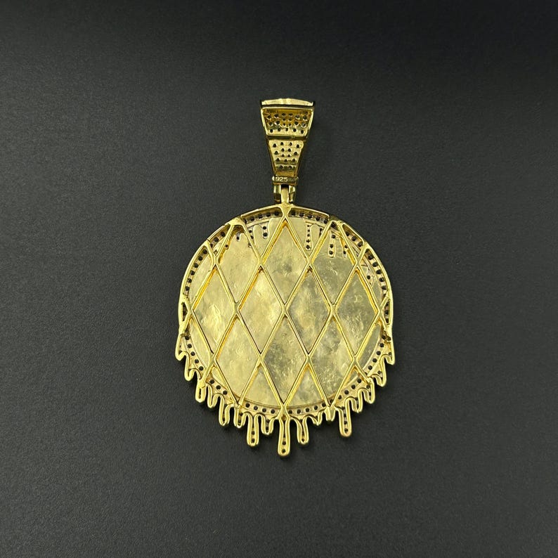 Op de afbeelding: Een goudkleurige hanger met een ruitvormig midden en een decoratieve, druipende rand. De hanger heeft een beugel voor een ketting en staat tegen een donkere achtergrond. Het sieraad is waarschijnlijk gemaakt van metaal en heeft een gedetailleerd ontwerp.