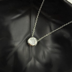 May include: A silver necklace with a round, clear gemstone pendant. The pendant is set in a bezel setting and hangs from a delicate chain. The necklace is displayed against a dark background.