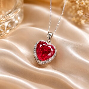 May include: A heart-shaped red gemstone pendant necklace featuring a halo of small white round stones set in a silver-toned metal frame. The pendant hangs from a delicate silver-toned chain, showcasing a faceted surface that reflects light.