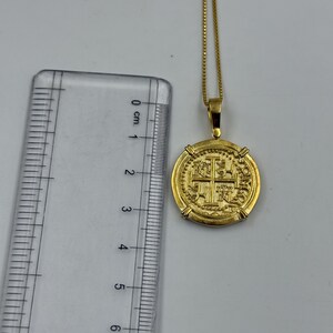 May include: A gold-colored coin pendant necklace. The pendant is round with an embossed design featuring a cross and other symbols. The chain is a delicate gold color. The pendant measures approximately 1 inch in diameter.