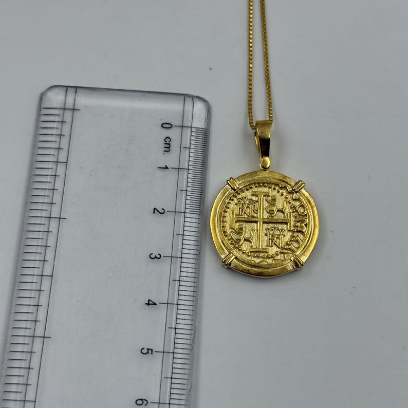 May include: A gold-colored coin pendant necklace. The pendant is round with an embossed design featuring a cross and other symbols. The chain is a delicate gold color. The pendant measures approximately 1 inch in diameter.
