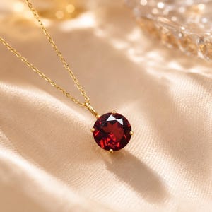 May include: A gold chain necklace featuring a round, faceted deep red garnet gemstone pendant held in a four-prong setting. The pendant hangs from a delicate gold link chain, showcasing the vibrant color and clarity of the stone.