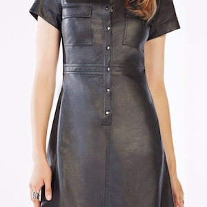 May include: A black short-sleeved shirt dress made of a smooth, leather-like material. The dress features a classic collar, a front button-down placket with metallic buttons, and two chest pockets with flaps. The silhouette is fitted at the waist.