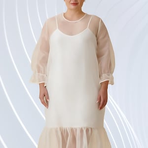 Puede incluir: Vestido color marfil con forro y sobrecapa transparente. El vestido presenta un escote redondo, mangas tres cuartos y un dobladillo con volantes. La modelo lleva sandalias blancas. El fondo tiene líneas blancas y azul claro.