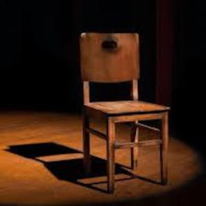 May include: A vintage wooden chair with a square seat and a rectangular backrest. The chair is made of light brown wood and has a simple, sturdy design. The chair is positioned on a wooden floor, with a dark background and a spotlight illuminating the chair.