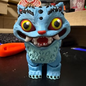 May include: A 3D-printed blue cat-like figure with large yellow eyes, red accents, and sharp teeth. The figure has black details on its head and paws, and a textured appearance. The cat is standing on a dark surface.