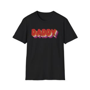 Puede incluir: Camiseta negra con la palabra "DADDY" en un diseño retro en capas. El texto utiliza tonos de rojo, naranja, blanco y morado, creando un efecto 3D. La camiseta tiene un cuello redondo clásico y mangas cortas.