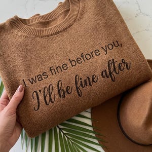 May include: A brown sweater with the text "I was fine before you, I'll be fine after" in black script. A brown hat and a green palm leaf are next to the sweater. The sweater is folded and held by a hand.