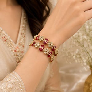 May include: Two gold bangles featuring oval-shaped red stones and small clear stones arranged in floral patterns. The bangles have a metallic gold finish and are designed with a repeating motif of red gems surrounded by clusters of tiny white stones.