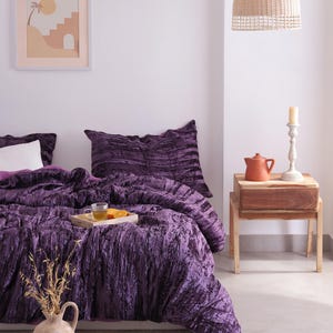 May include: A bedroom with a bed covered in a deep purple, textured duvet and pillows. A wooden tray holds a cup of tea and orange slices. A framed print hangs on the wall, and a woven pendant light hangs from the ceiling.