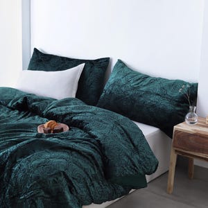 Puede incluir: Una cama con un edredón de terciopelo verde oscuro y fundas de almohada a juego. Una almohada blanca se coloca frente a las almohadas verde oscuro. Una mesita de noche de madera tiene un jarrón con flores y velas.