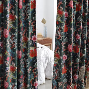 Puede incluir: Cortinas verde oscuro con estampado floral en rojo y azul. Las cortinas presentan un diseño repetido de flores y hojas. La imagen muestra las cortinas parcialmente abiertas, revelando un dormitorio con un edredón blanco y una mesita de noche de madera.