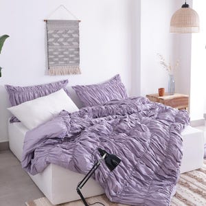 Puede incluir: Una escena de dormitorio con una cama con un edred&oacute;n fruncido lavanda y fundas de almohada a juego. Una almohada blanca contrasta con la ropa de cama lavanda. Una l&aacute;mpara de escritorio negra descansa sobre libros. Un tapiz tejido y una alfombra a&ntilde;aden textura.