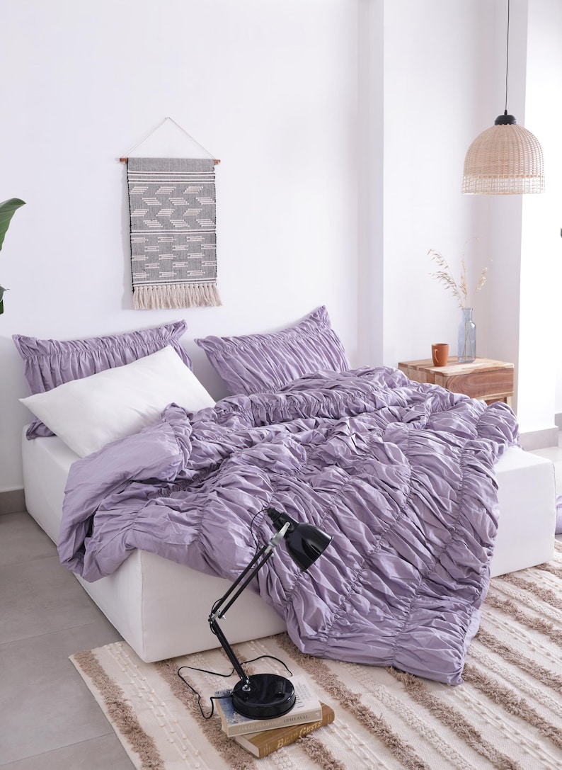 Puede incluir: Una escena de dormitorio con una cama con un edred&oacute;n fruncido lavanda y fundas de almohada a juego. Una almohada blanca contrasta con la ropa de cama lavanda. Una l&aacute;mpara de escritorio negra descansa sobre libros. Un tapiz tejido y una alfombra a&ntilde;aden textura.