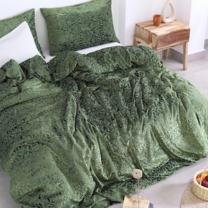 Può includere: Un letto con un piumino di velluto verde e federe abbinate, con un motivo ornato. È visibile anche un cuscino bianco. Un comodino in legno e un vaso con piante essiccate sono accanto al letto.